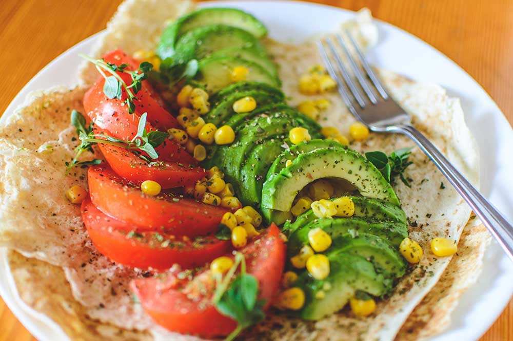 A plate featuring a single tortilla topped with wedges of tomatoes and avocados along with corn and a sprinkling of spices. A fork lies next to the food.