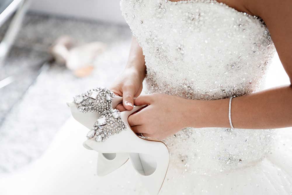 Close-up view of the torso of a woman in an embroidered wedding gown. She holds a pair of white high heels in her hands. The backs of the shoes are elaborately encrusted in sparkling crystal rhinestones in a variety of shapes and sizes.