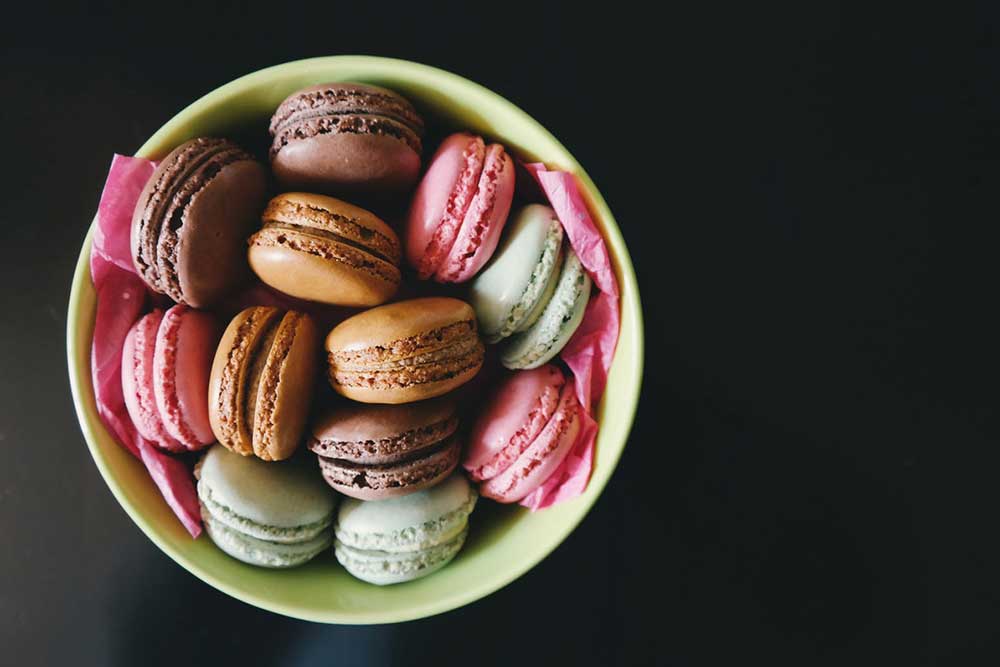 Overhead view of a green bowl lined with pink tissue paper and filled with a dozen macarons in varying shades of pink, green, and brown.