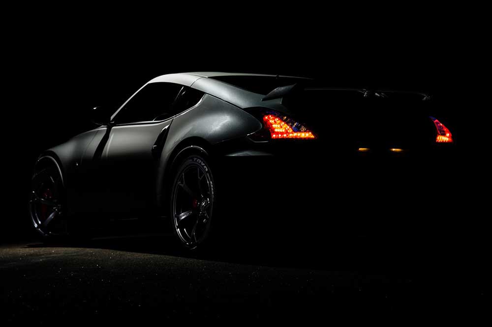 View of a two-door sports car from behind. It's dramatically lit in a way that showcases the vehicle's slick design and matte black paint job while still casting the entire image in a dark, moody light. The only color in the image is the illuminated red brake lights of the car.