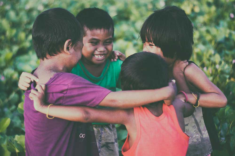 Four small children in t-shirts and tank tops stand outside in a circle with their arms thrown around each other and excitedly embrace.