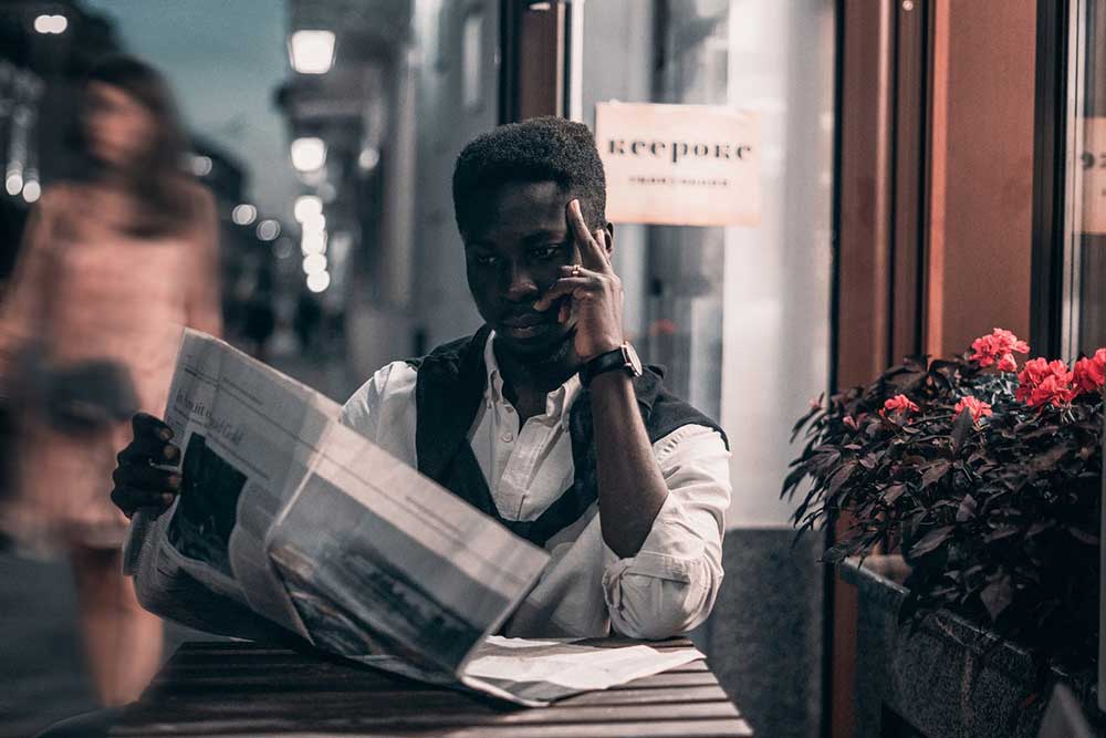 Young man reads a newspaper at a table outside a cafe window in the early evening. He wears a white oxford shirt with the sleeves rolled to his elbows, a dark cardigan casually draped around his shoulders, and a sleek leather-banded watch.