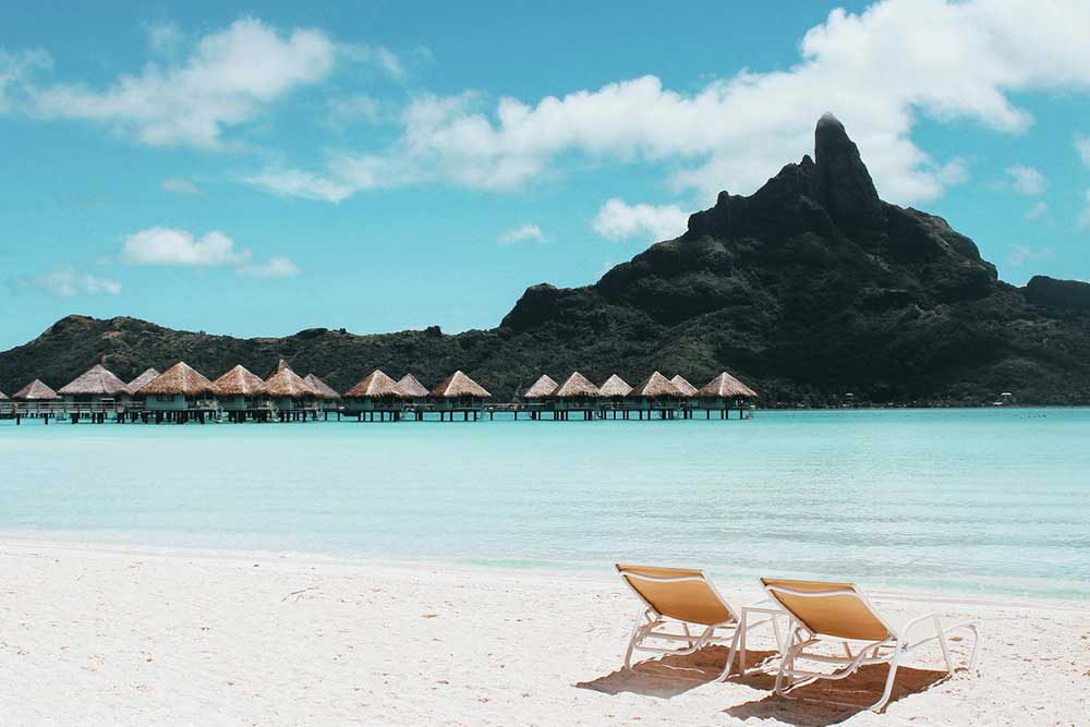 Two tan pool chairs sit on a white sand beach next to a crystal blue ocean. Beach cottages sit on stilts over the water and a tree-covered mountain peak is just beyond them in the distance. The sun shines brightly overhead and the sky is a brilliant blue.