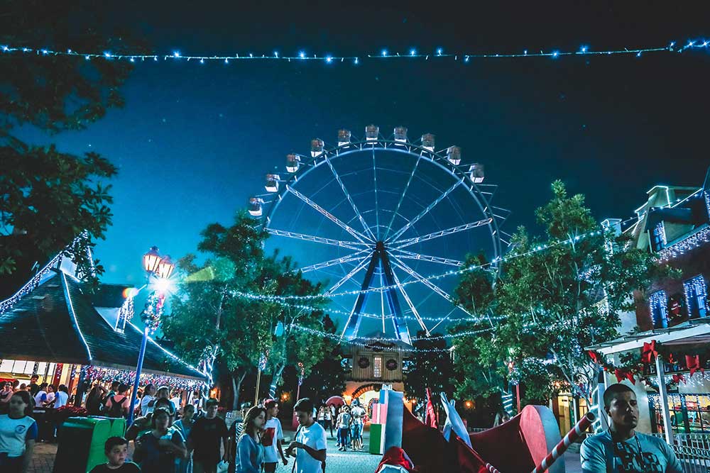 A Ferris Wheel lit up under a starry night sky at a carnival crowded with people and vendor stalls. Strands of string lights crisscross above where they walk.