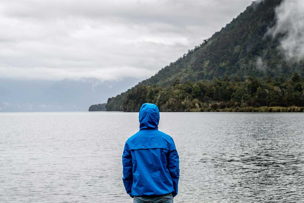 Person in a blue windbreaker jacket looks out over a large lake bordered by a steep hillside covered in trees. The word “peace” is ghosted over the image in large, bold letters.