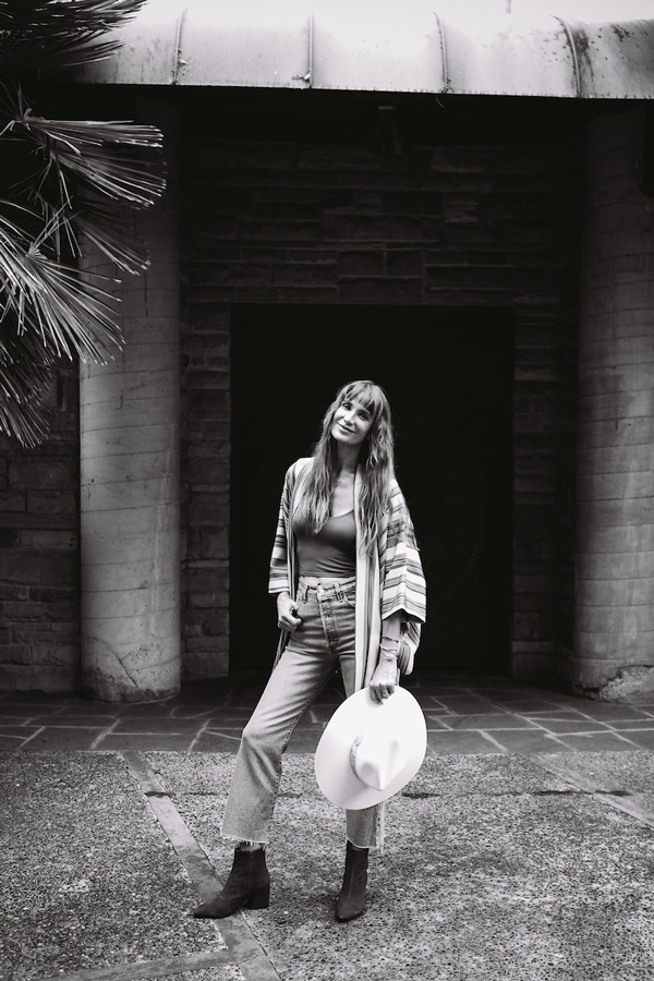 Young woman in high-waisted jeans and patterned cardigan holding a wide-brimmed hat, standing in front of a stone building entrance.