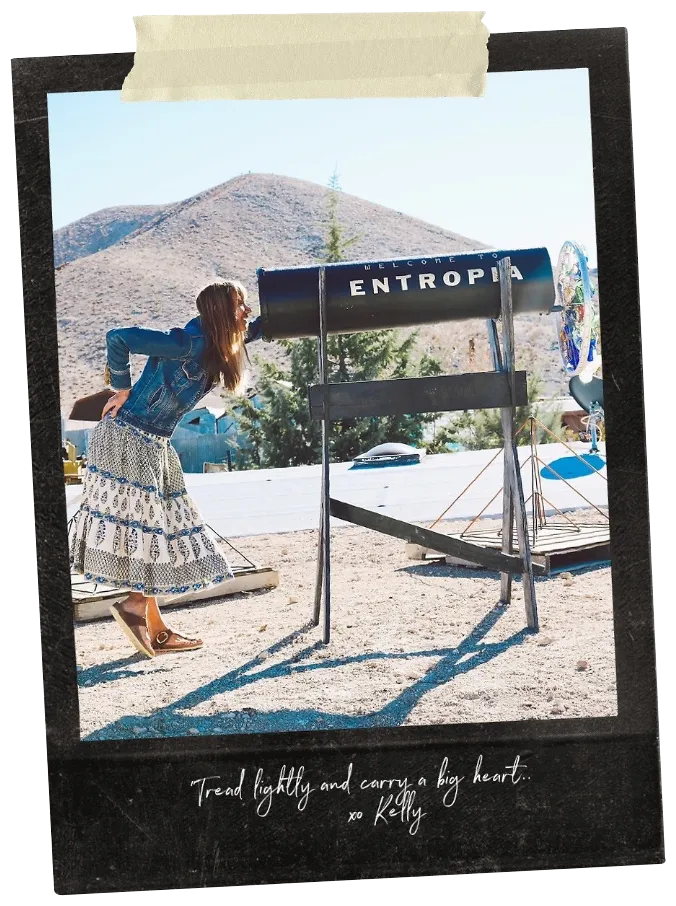 Woman in a patterned skirt and denim jacket looking through a large black telescope labeled 'ENTROPIA' outdoors with mountains in the background.