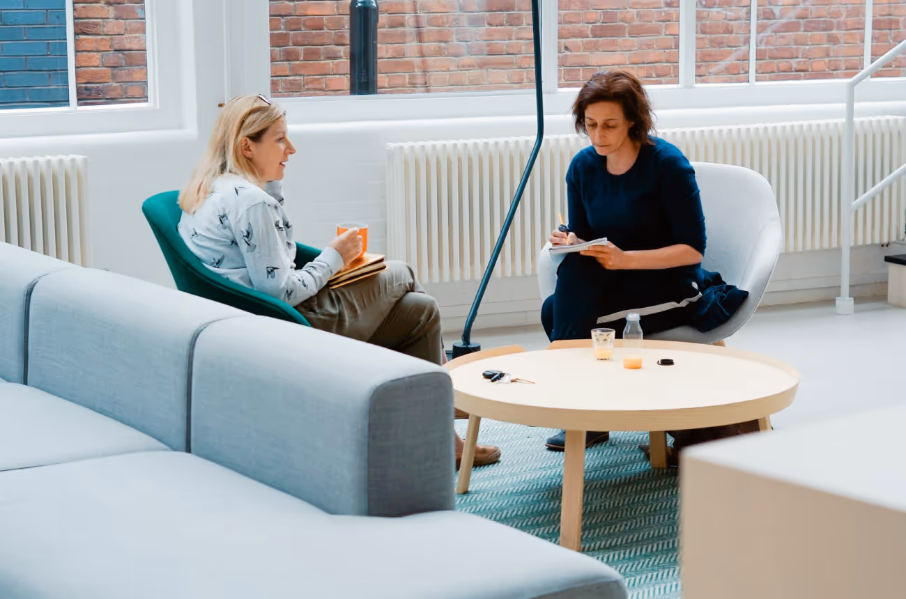 Two women talk in a modern lounge; one takes notes, the other holds a mug and folders for employee engagement.