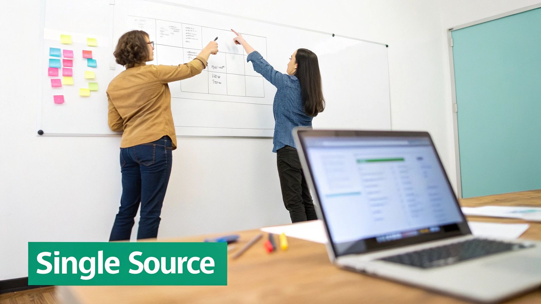 Two women collaborate on a whiteboard, writing and pointing, with sticky notes nearby in an office setting.