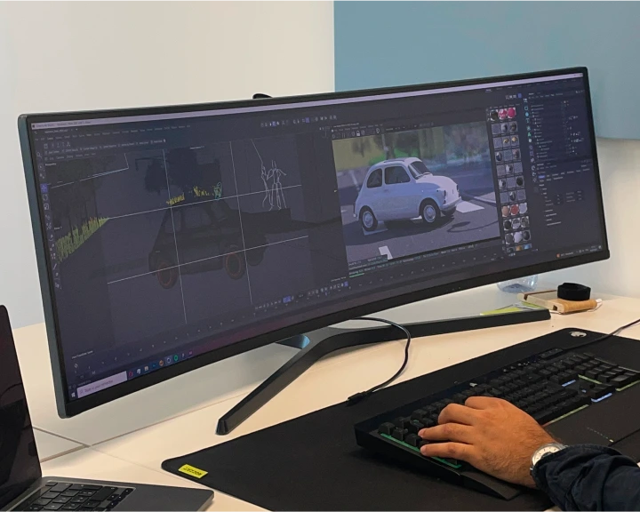 Écran ultra-large affichant un logiciel de modélisation 3D avec une voiture blanche en création, main droite d'une personne utilisant un clavier visible.