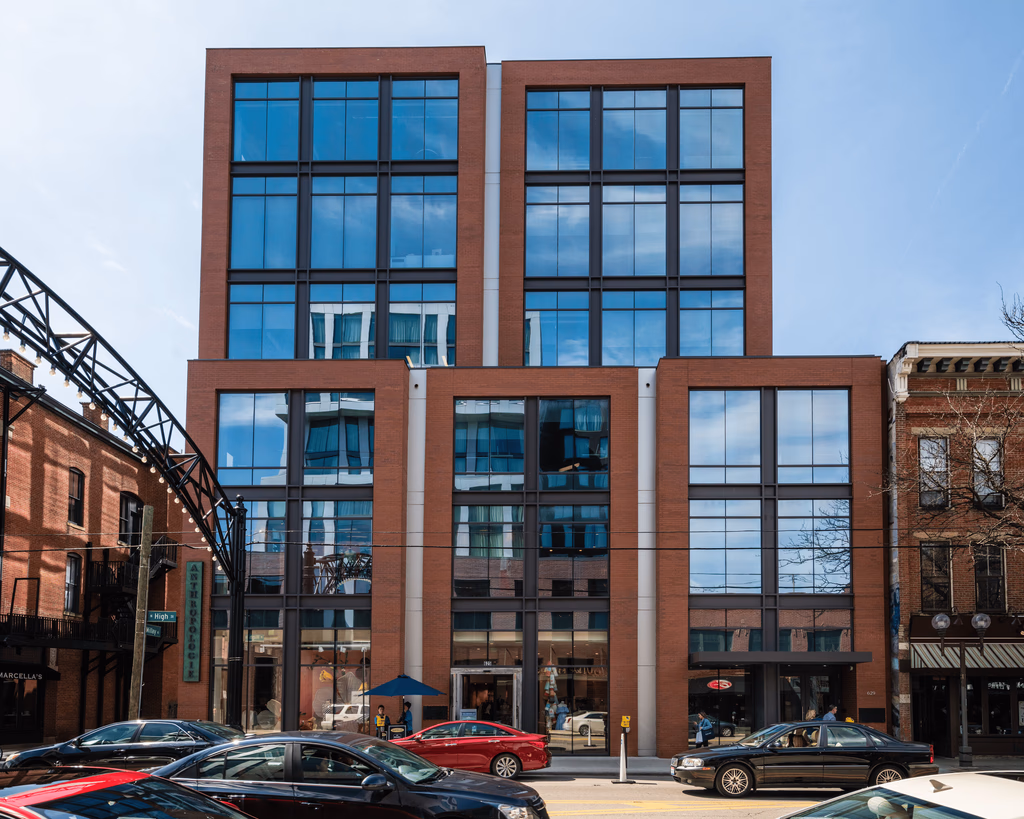 A modern brick building with large reflective windows, under a clear blue sky. Cars pass by on the street in front. The scene feels urban and bustling.