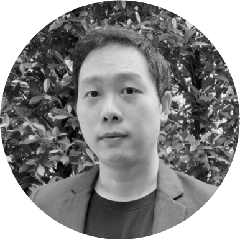 Portrait of a man with short hair wearing a dark blazer and t-shirt, standing in front of leafy bushes.