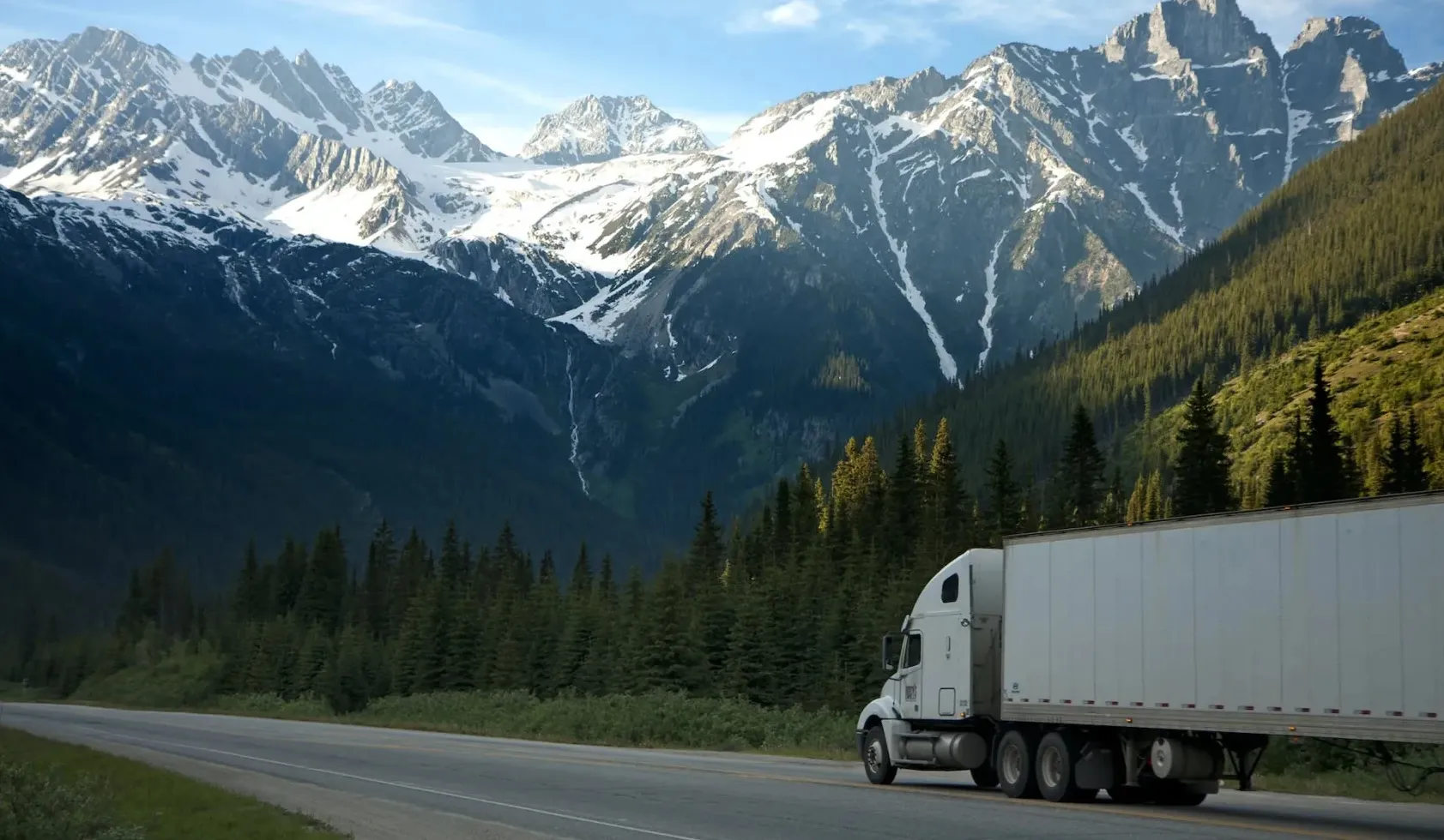 White semi-truck driving on road beside dense forest with snow-capped mountains in the background.
