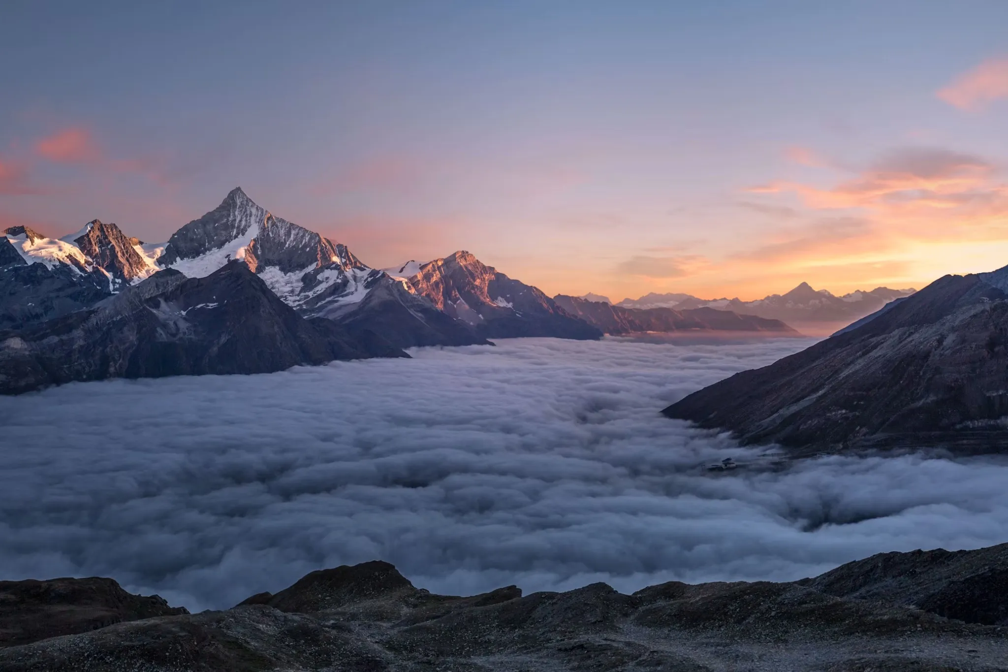 Mountain range at sunrise with snow-capped peaks above a thick sea of clouds in a valley.