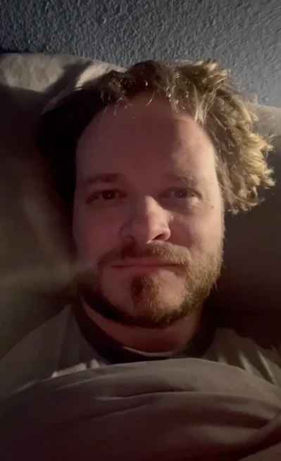 Man with light brown hair and beard lying in bed under a blanket looking at the camera.