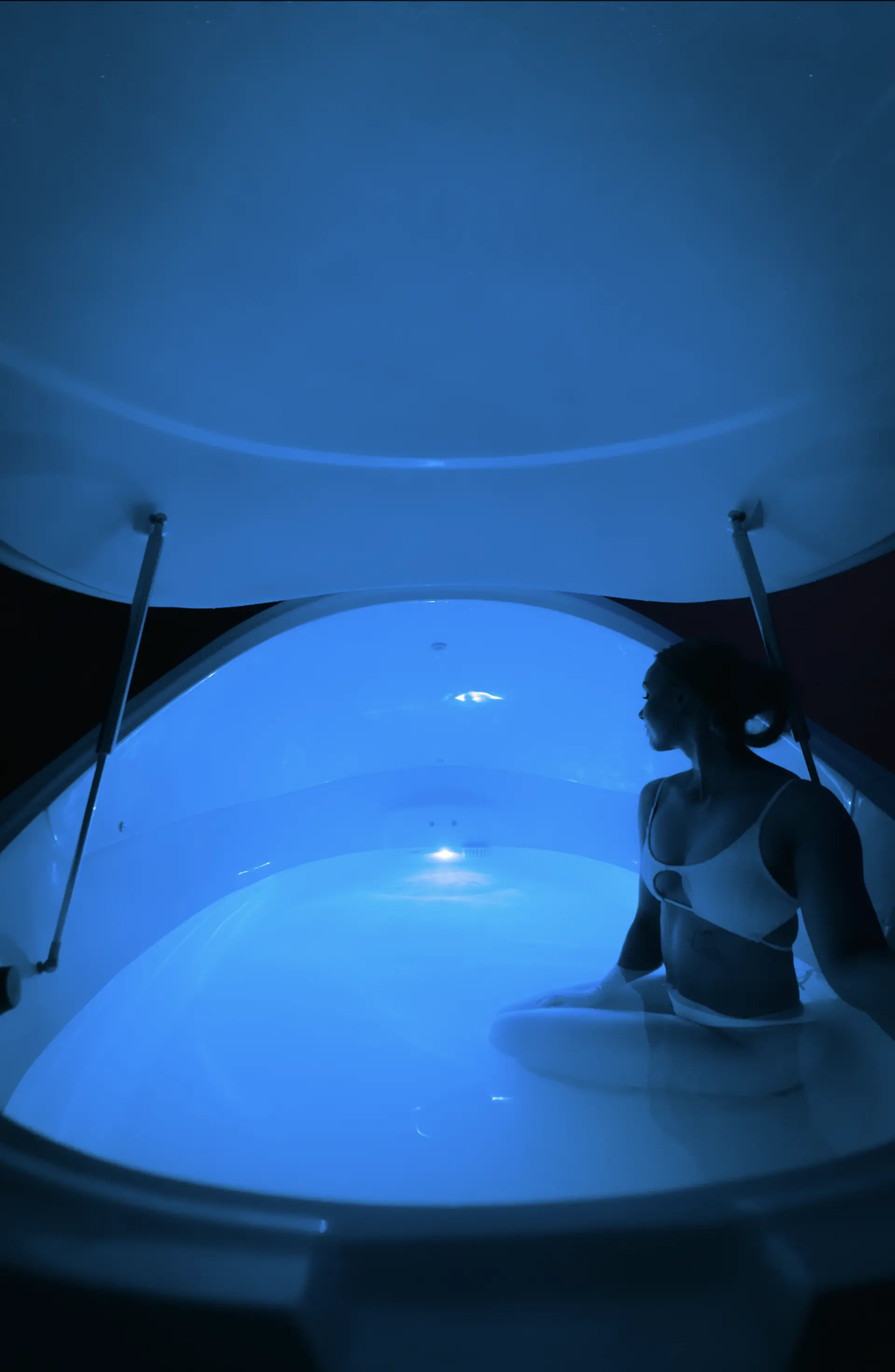 Nainen istuu rauhallisesti kelluvassa kylpytynnyrissä sinisessä valaistuksessa.