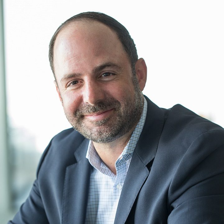 Professional headshot of bearded man in dark blazer and checkered shirt smiling