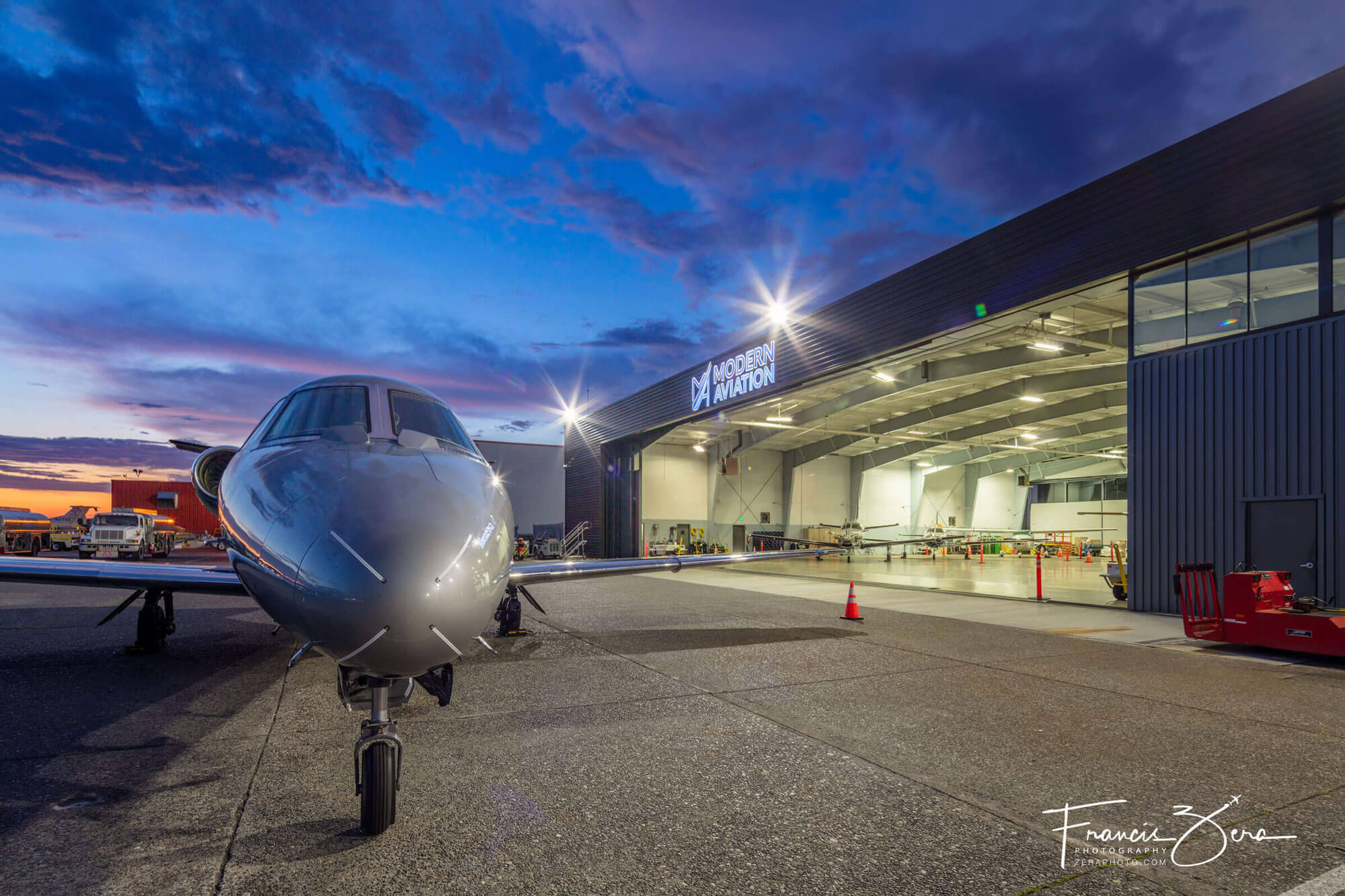 A twilight photo of a private jet aircraft in front of a brightly lit hangar