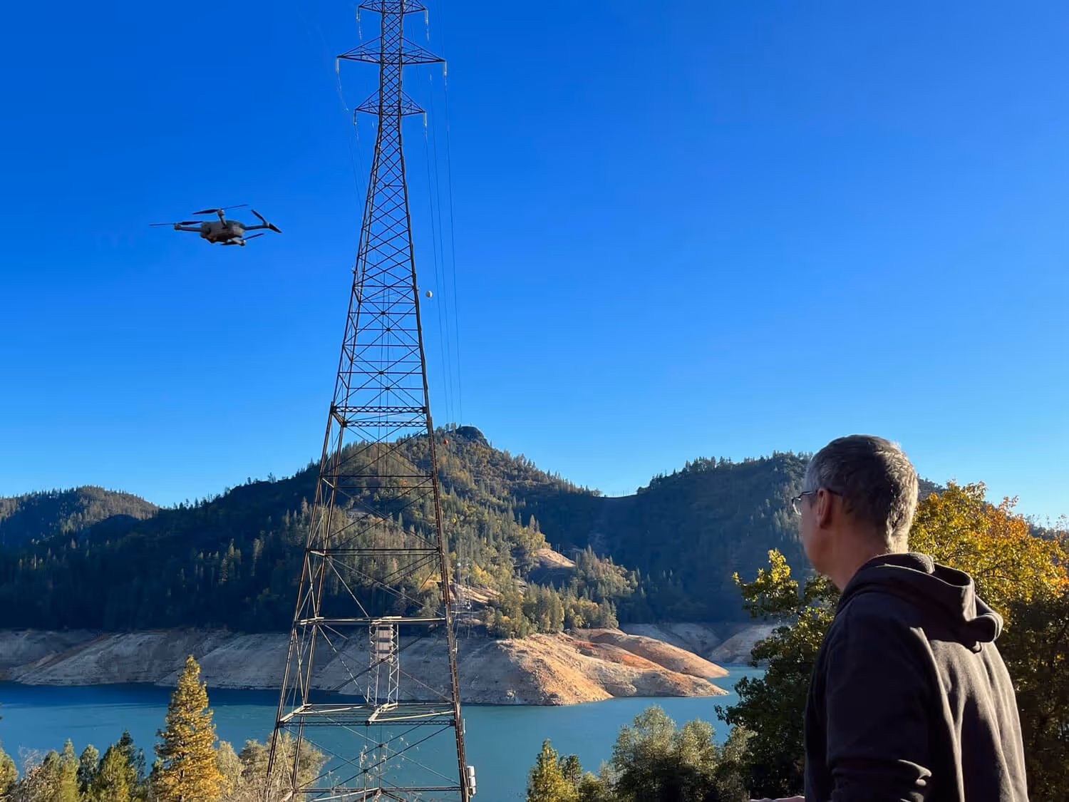 A commercial drone pilot flies a drone to inspect power pylons near a lake