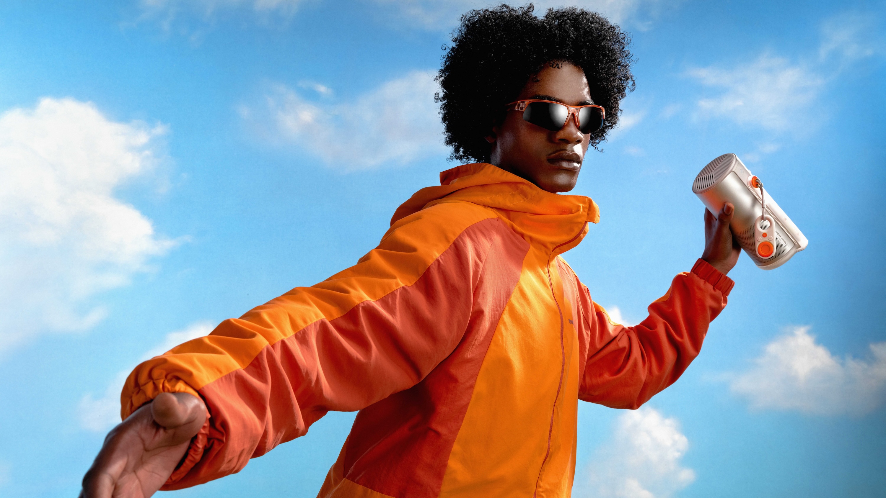 Young man wearing orange jacket and sunglasses holding a portable speaker against a blue sky with clouds.
