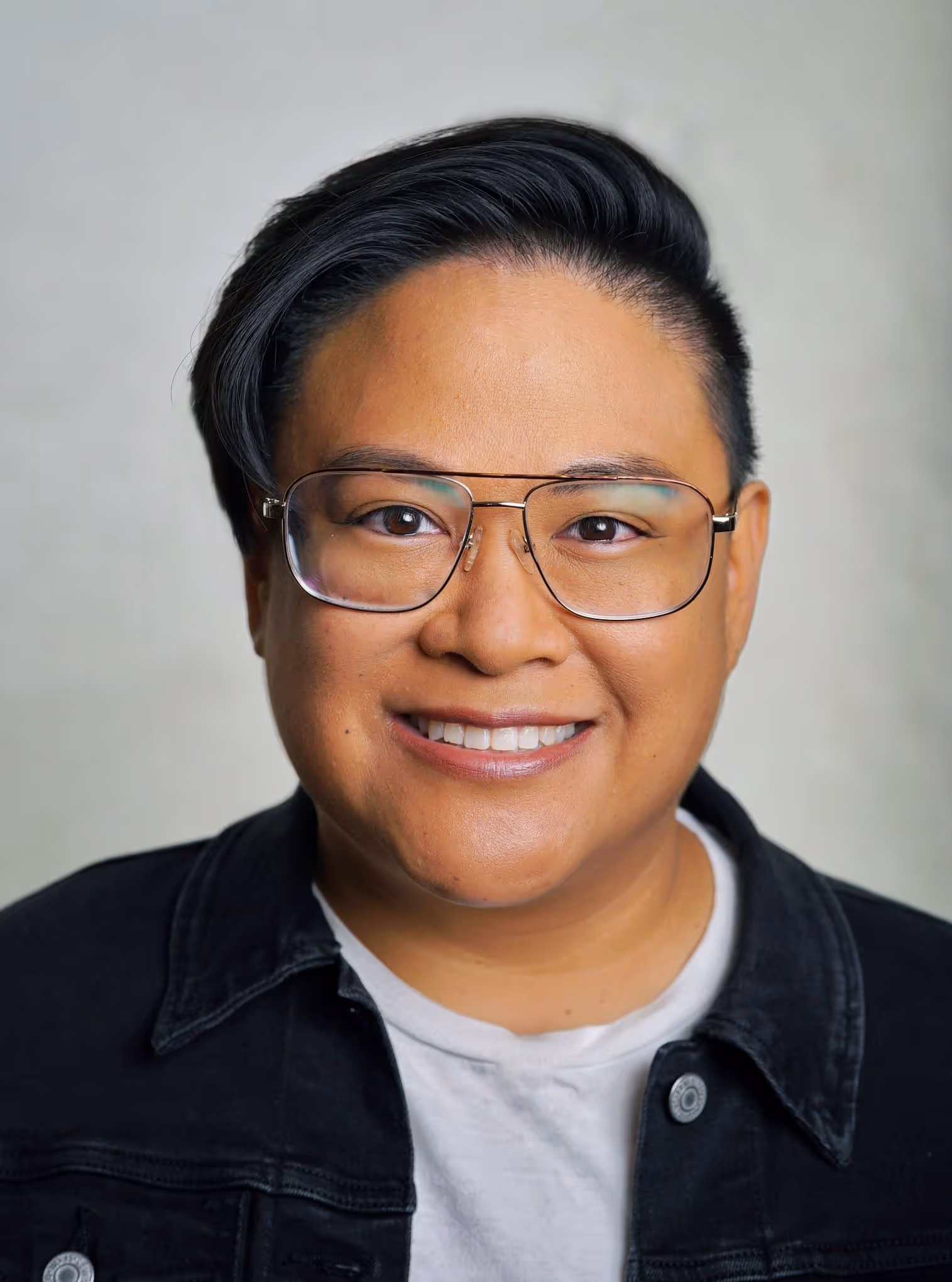 Smiling person wearing glasses and black denim jacket against neutral background