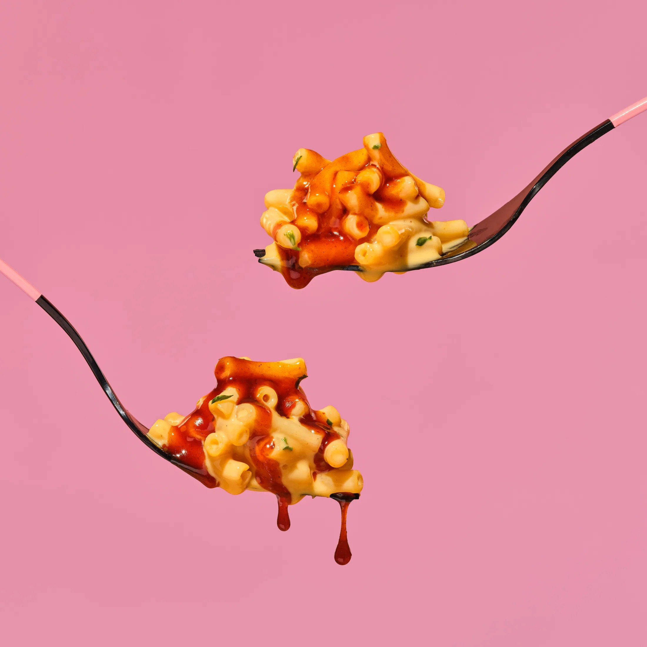 Two forks lifting carbonara pasta with sauce dripping against a pink backdrop