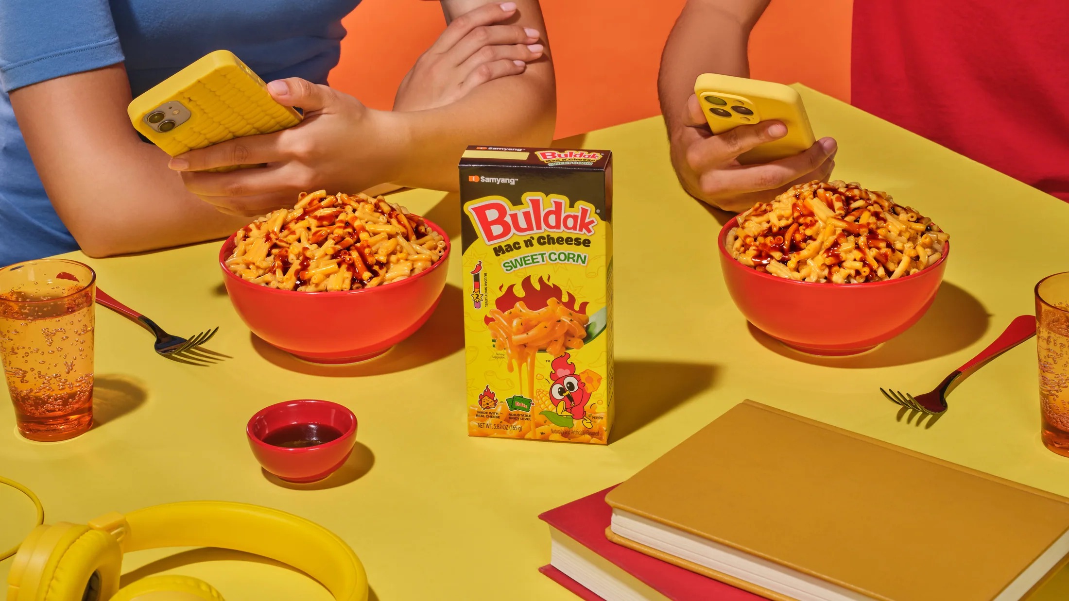 Two people on their phones with bowls of sweet corn pasta and a Buldak box