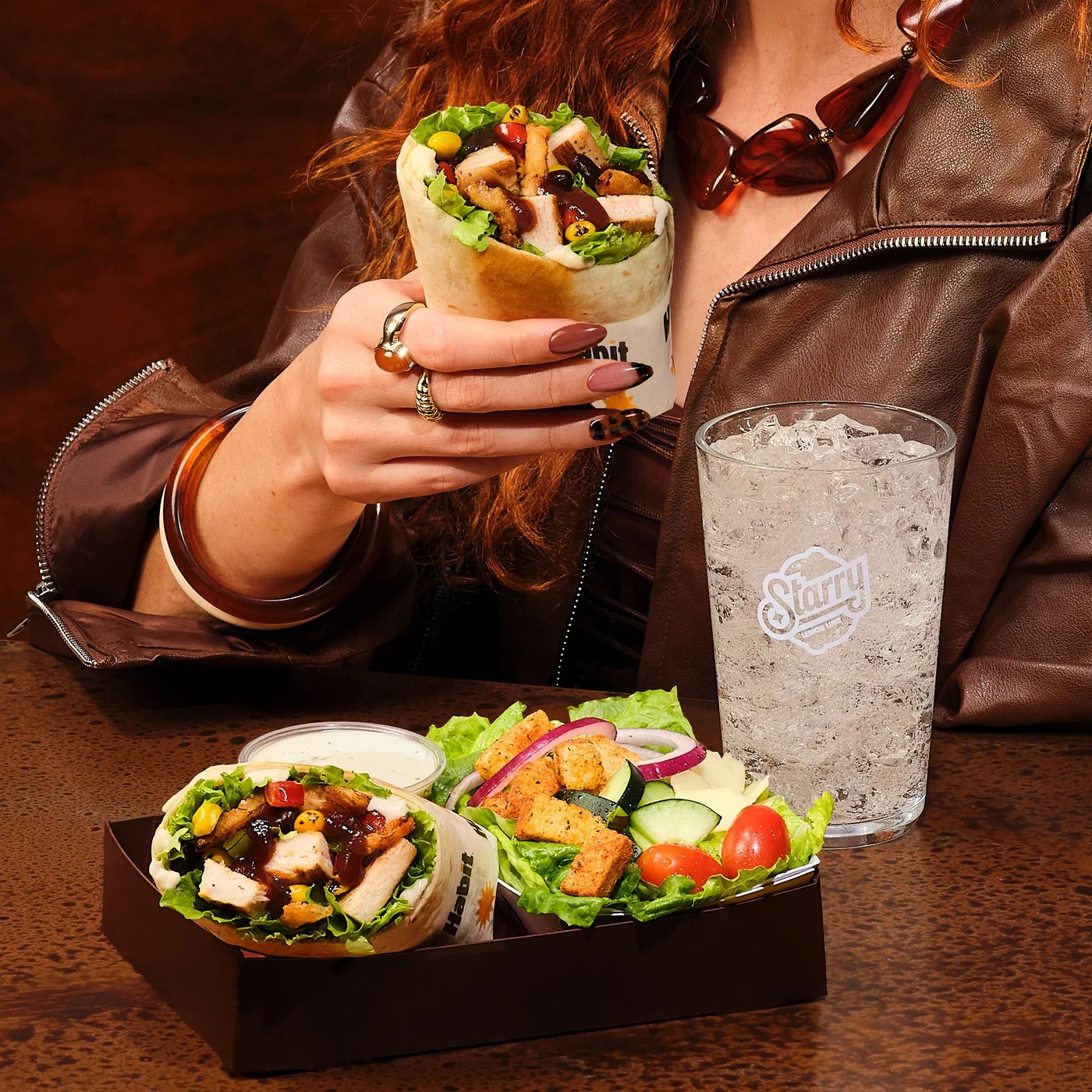 Person in a leather jacket holding a wrap beside two salads