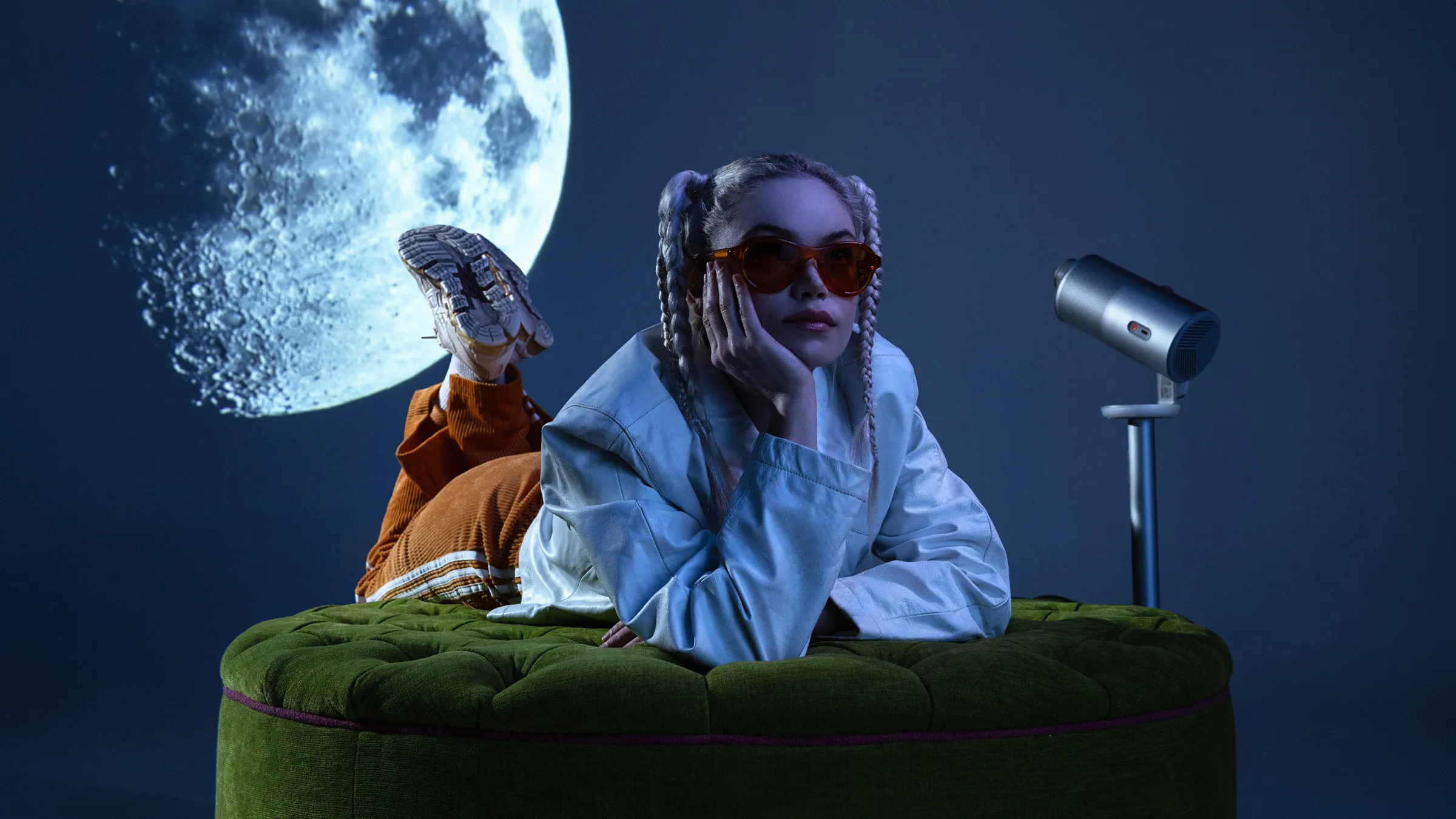 Person in red sunglasses lying on a green velvet ottoman beside a projector, a full moon projected on the wall behind
