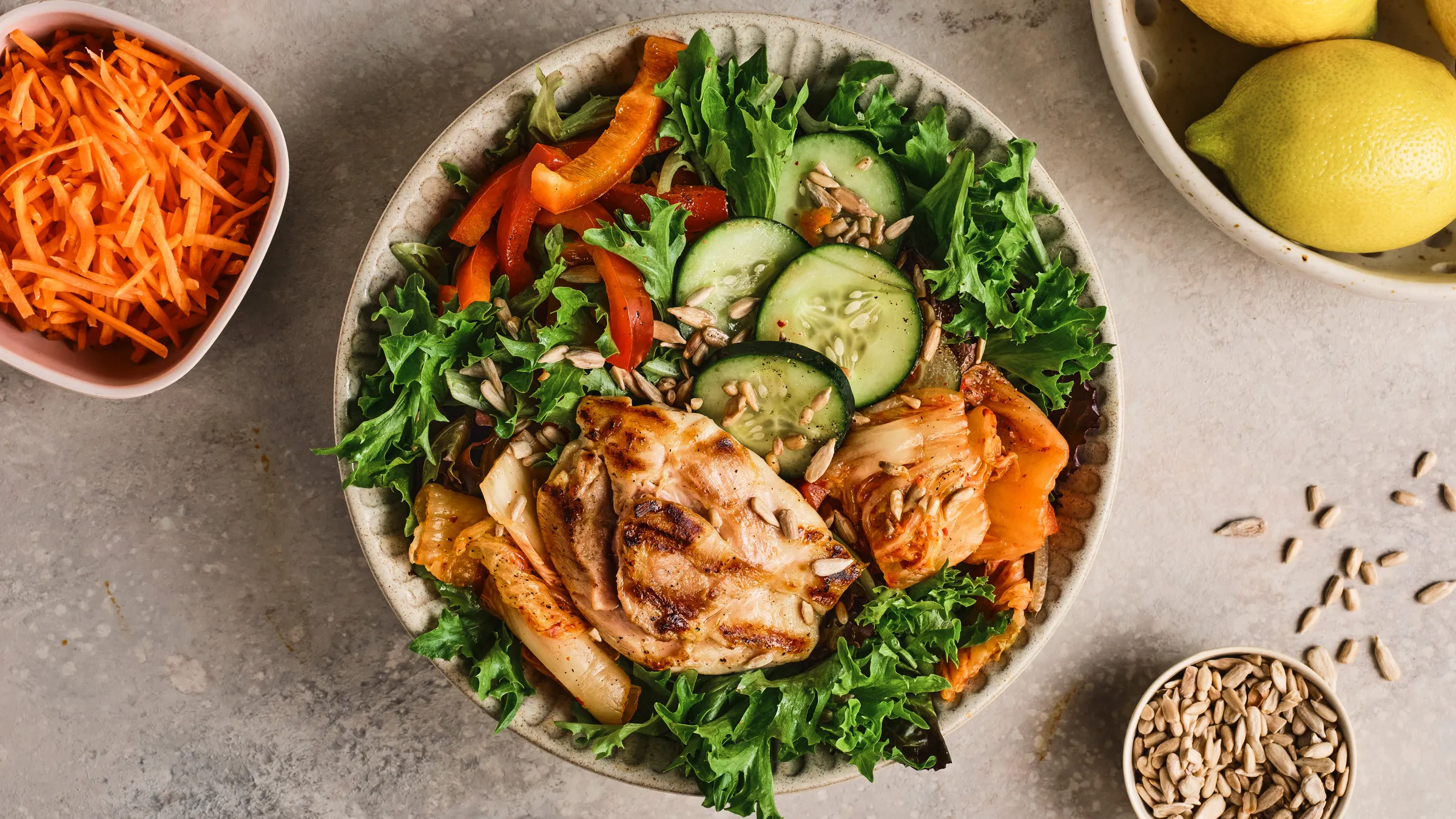 Overhead salad of grilled chicken thigh with kale, cucumber, peppers and sunflower seeds, beside grated carrot and lemons