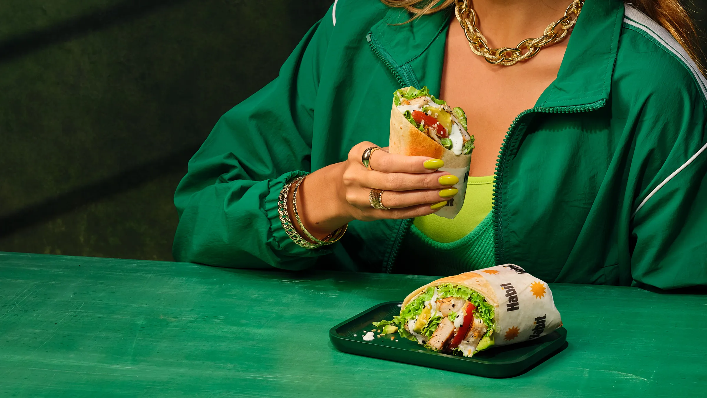 Person in a green tracksuit holding a chicken avocado wrap at a green table, a second wrap on a tray in front