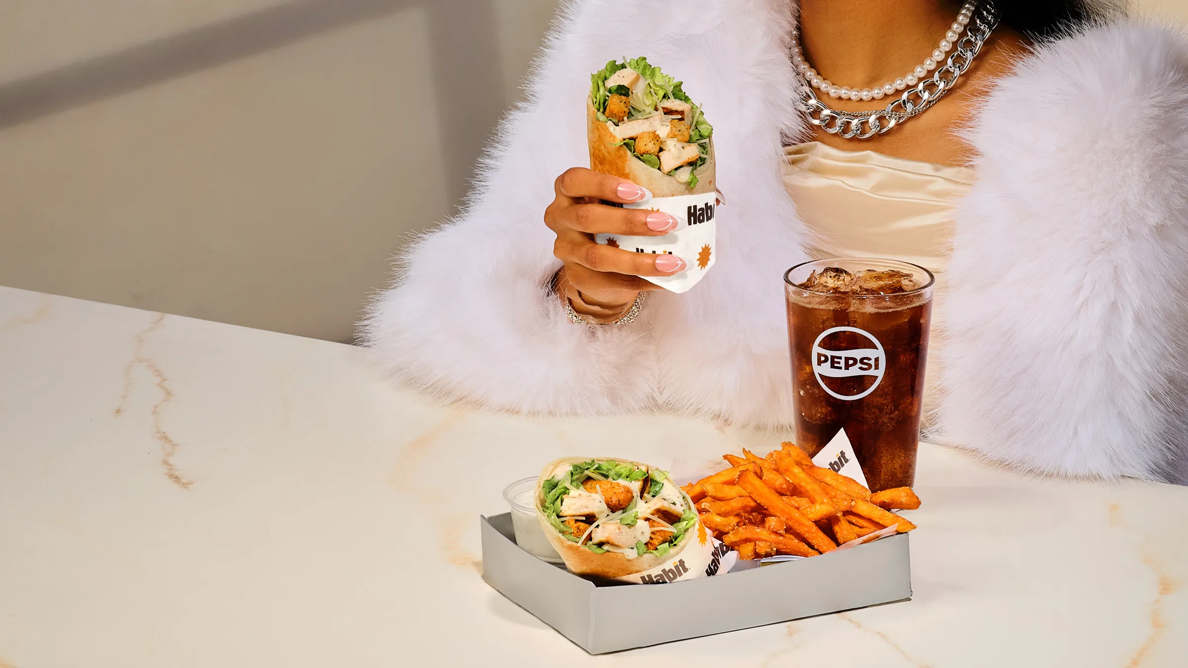 Person in a white fur coat holding a chicken caesar wrap, with a second wrap, sweet potato fries and a Pepsi on a marble table