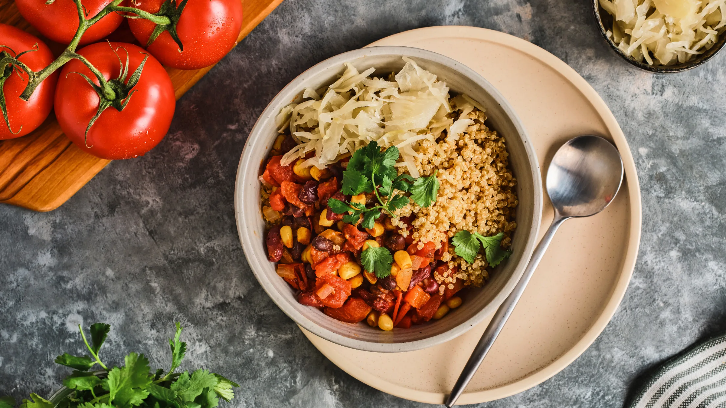 Overhead bowl of vegan chilli with corn, black beans and grains, topped with coriander, beside vine tomatoes