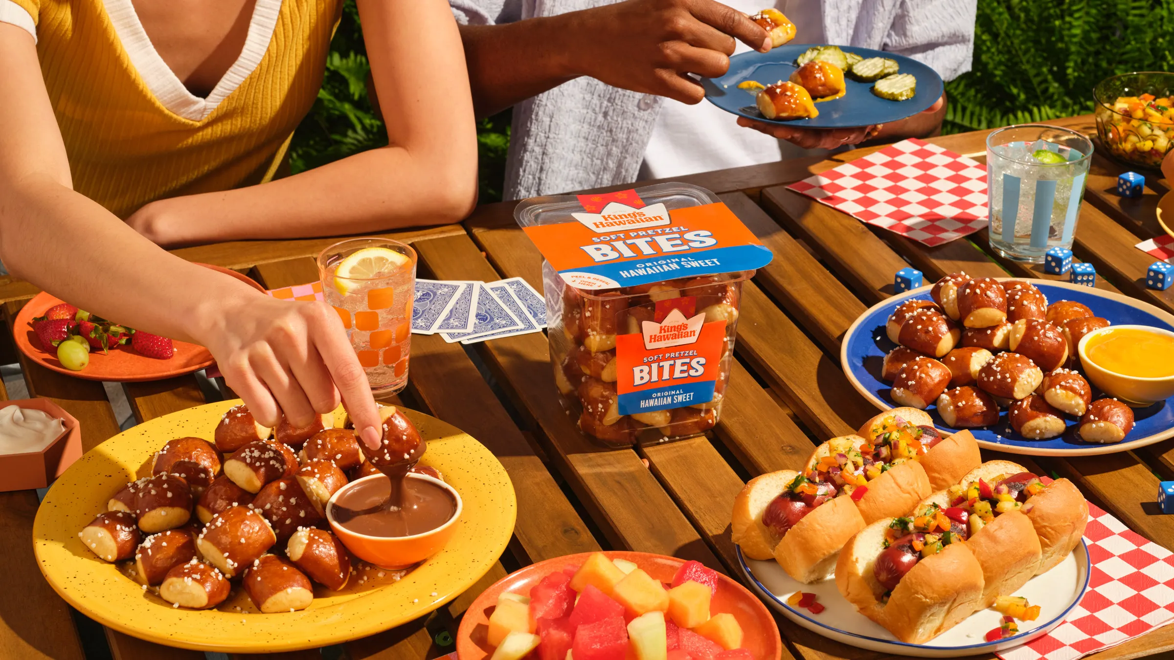Outdoor table of King's Hawaiian pretzel bites, sliders and fruit, with hands reaching in to share