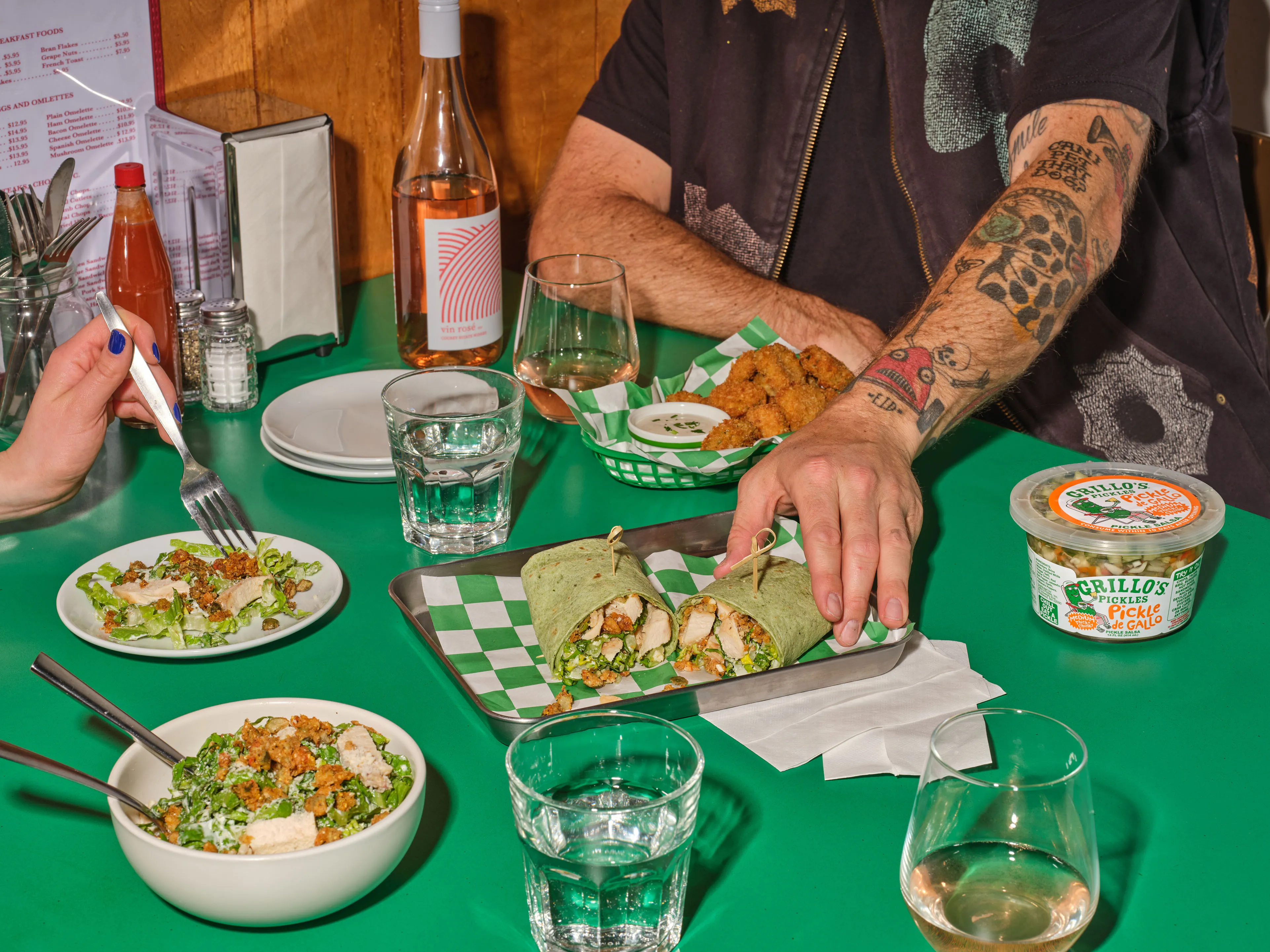 Two people at a green table sharing a grilled chicken caesar wrap, salads and a tub of Grillo's Pickles