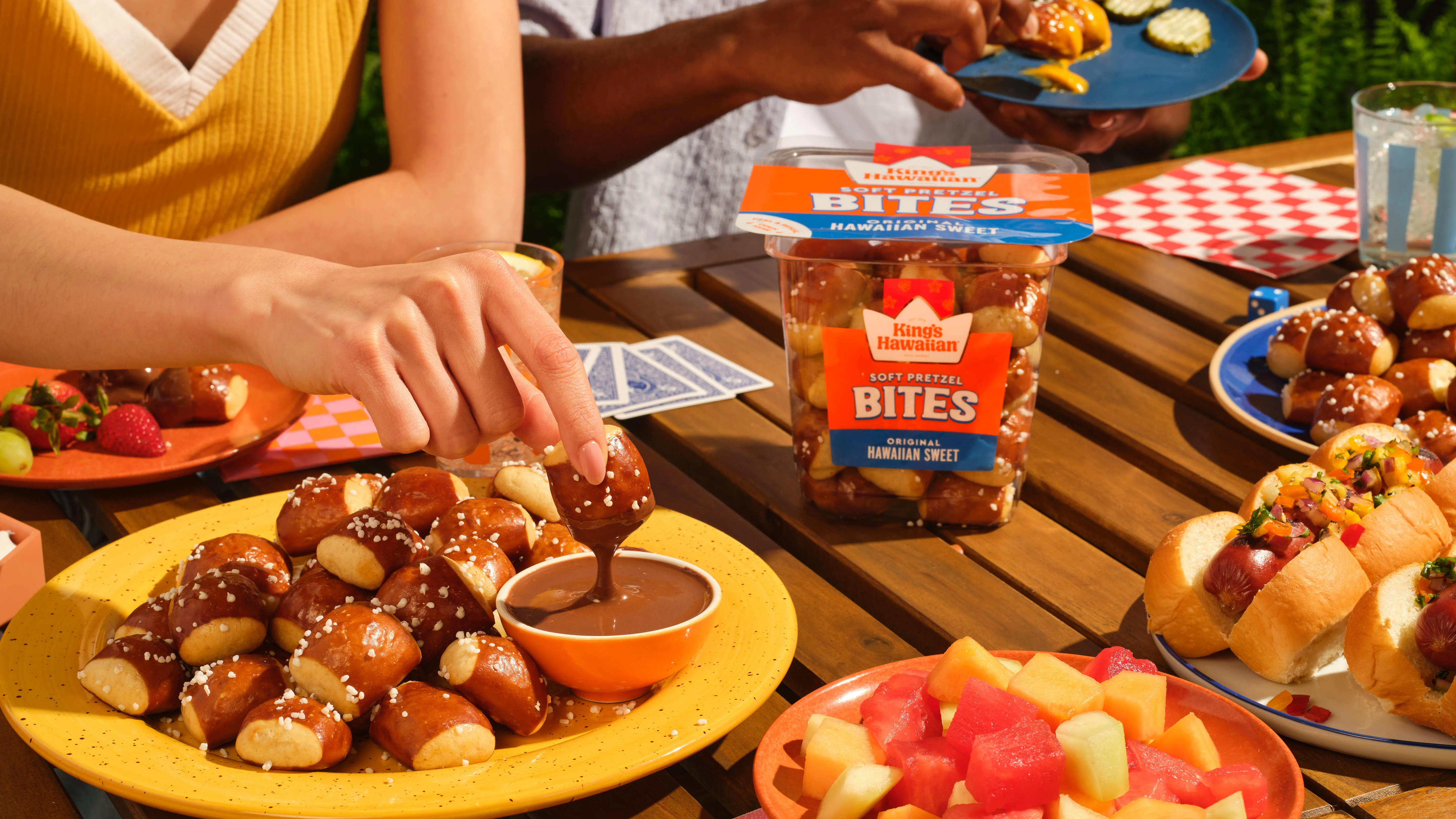 Hand dipping a King's Hawaiian soft pretzel bite into cheese sauce at a sunlit outdoor table