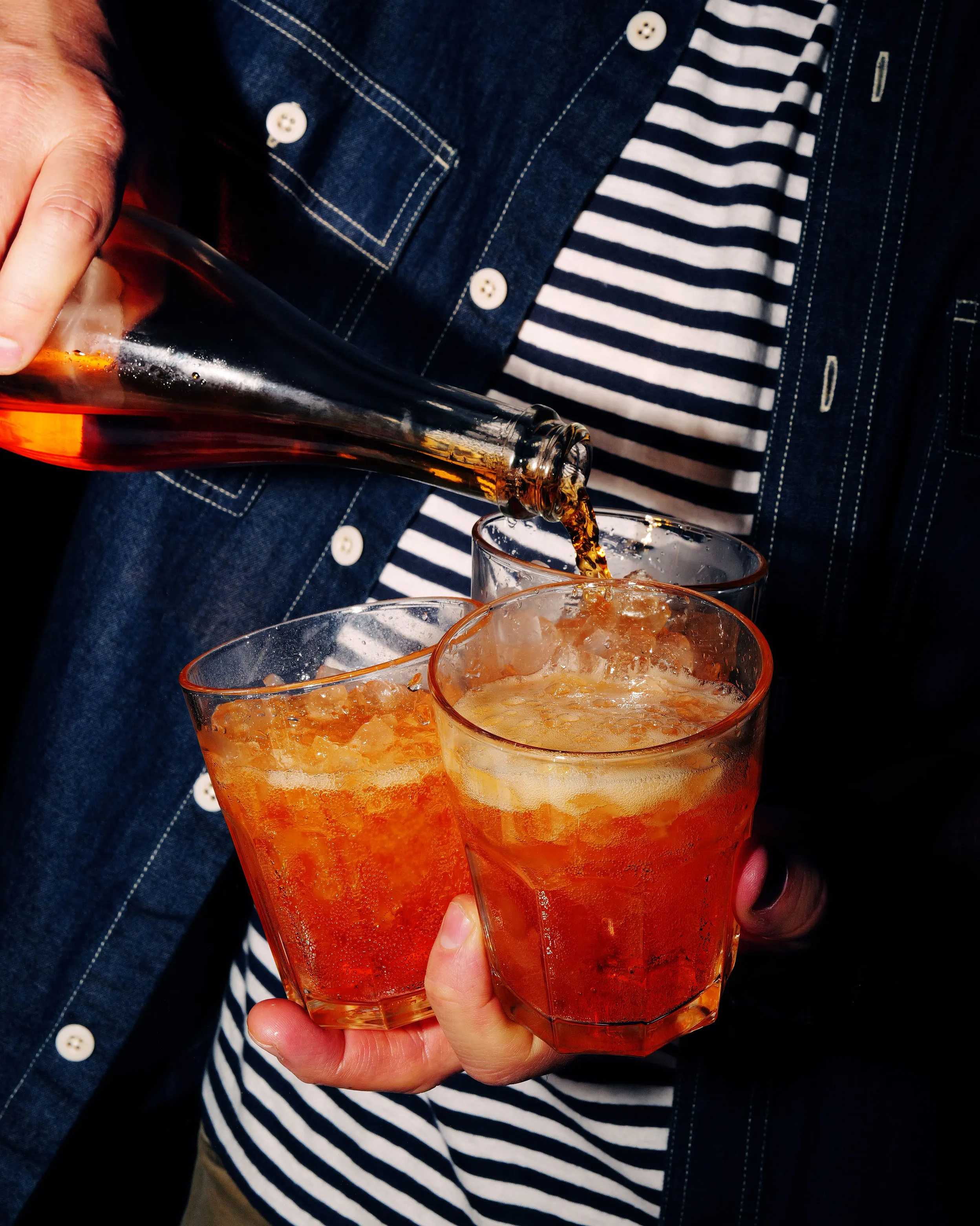 Person in a striped shirt pouring an amber cocktail from a bottle into three ice-filled glasses