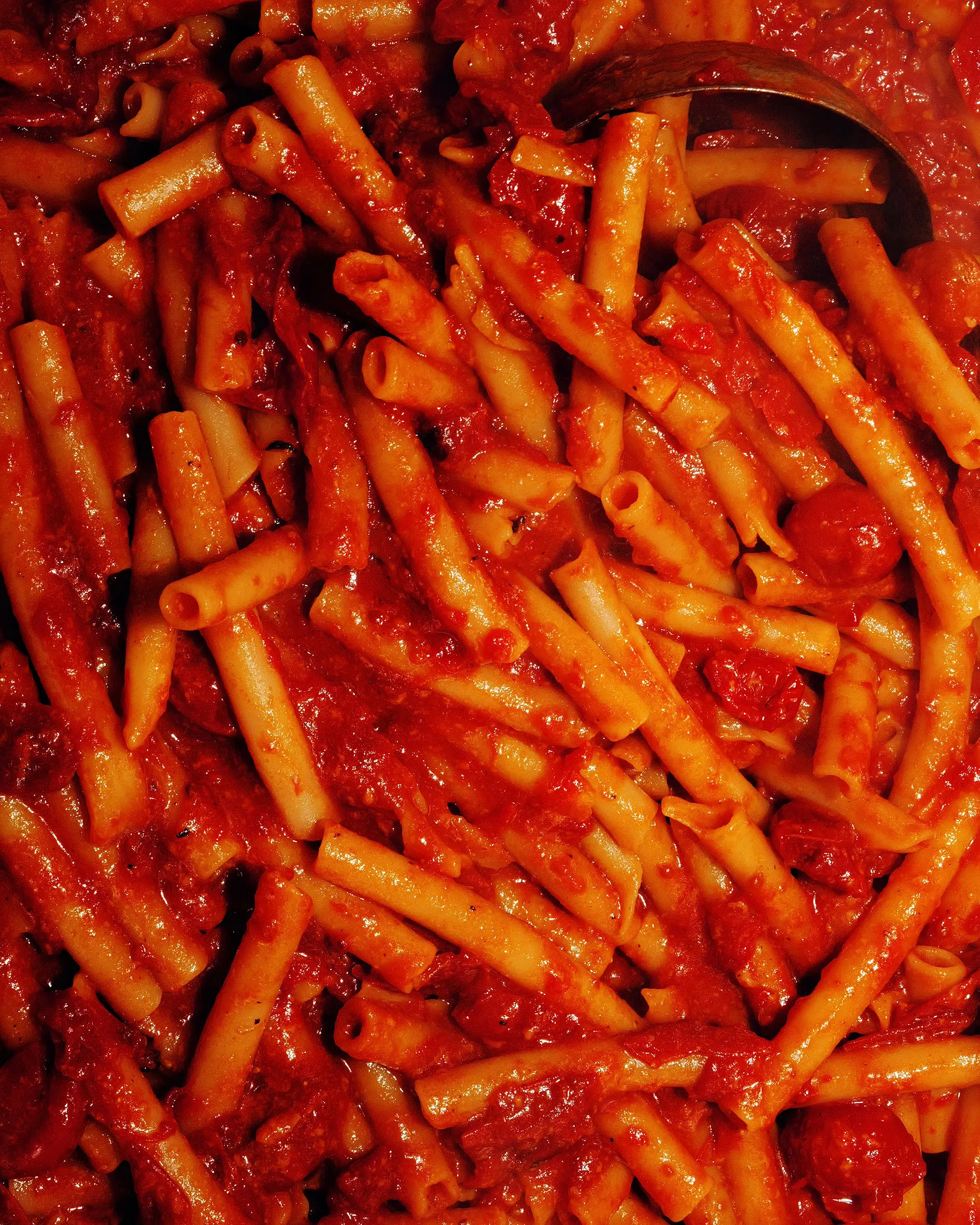 Overhead of a wide pan of baked ziti in tomato sauce with a serving spoon, on a wooden worktop