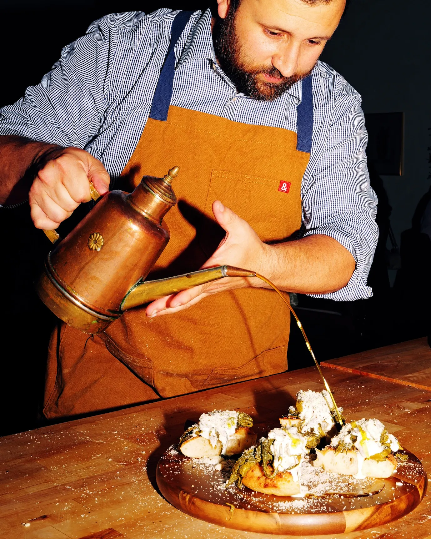 Chef pouring syrup from a copper pot over a dessert dusted with icing sugar on a wooden board