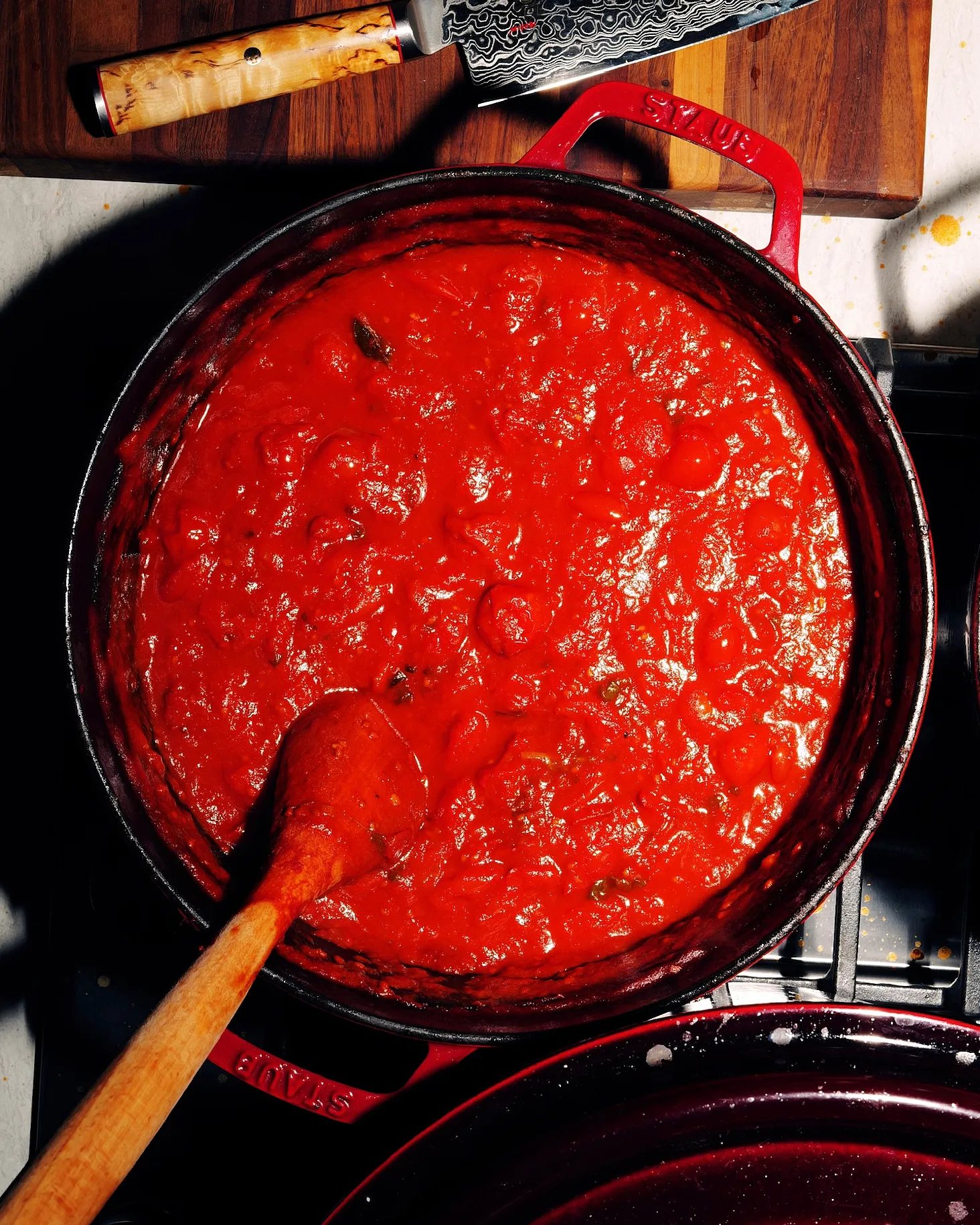 Overhead of a red enamel pan of tomato sauce on the hob, with a wooden spoon and a chef's knife alongside