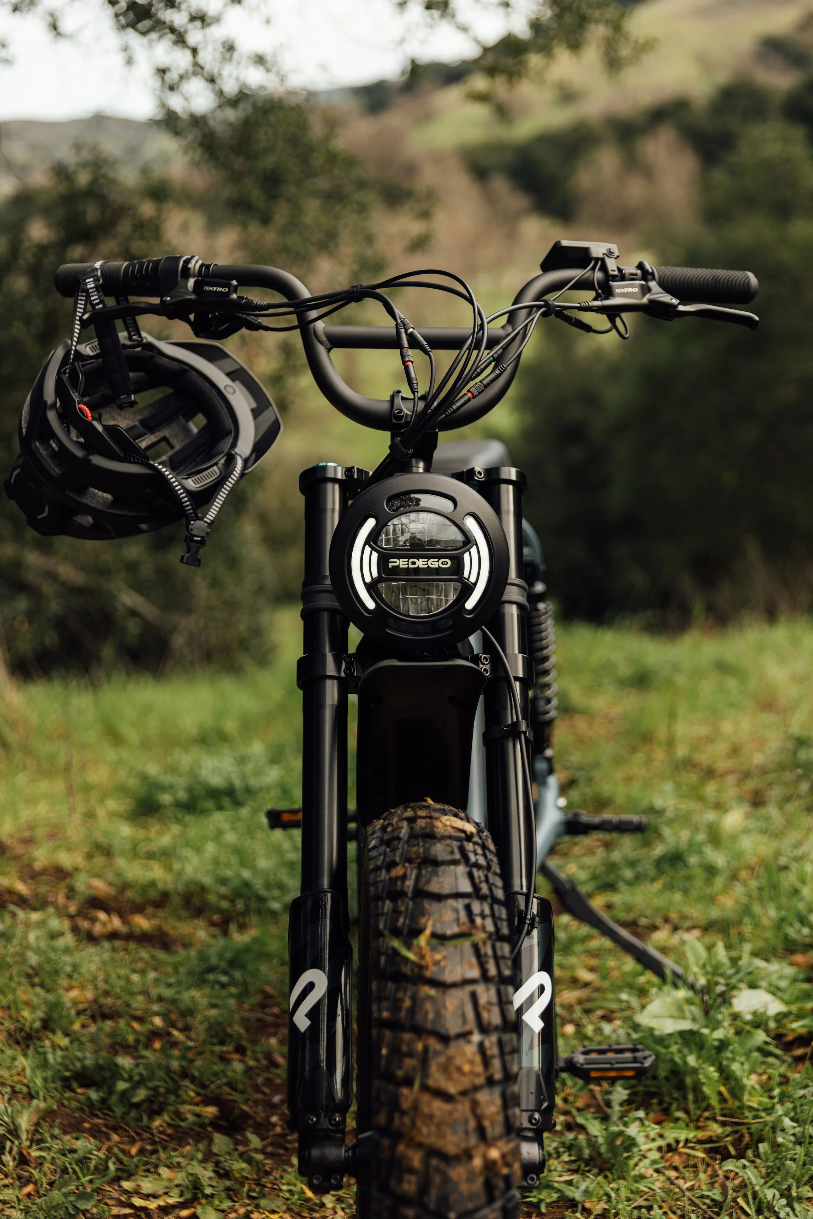 Front fork and headlight of a Pedego e-bike parked on grass, a helmet hanging from the handlebars