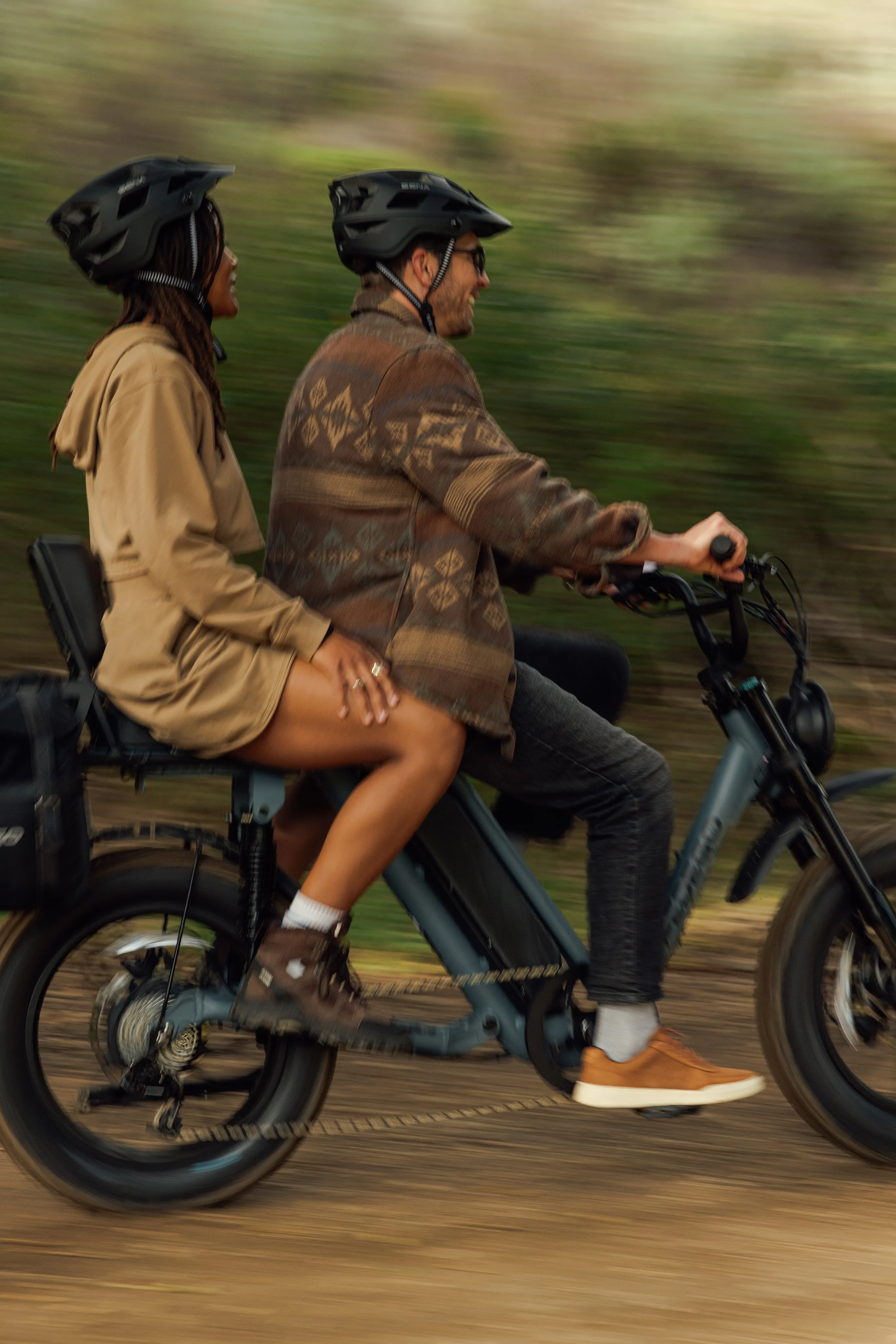 Two people riding a Pedego e-bike together along a trail, the background blurred with motion