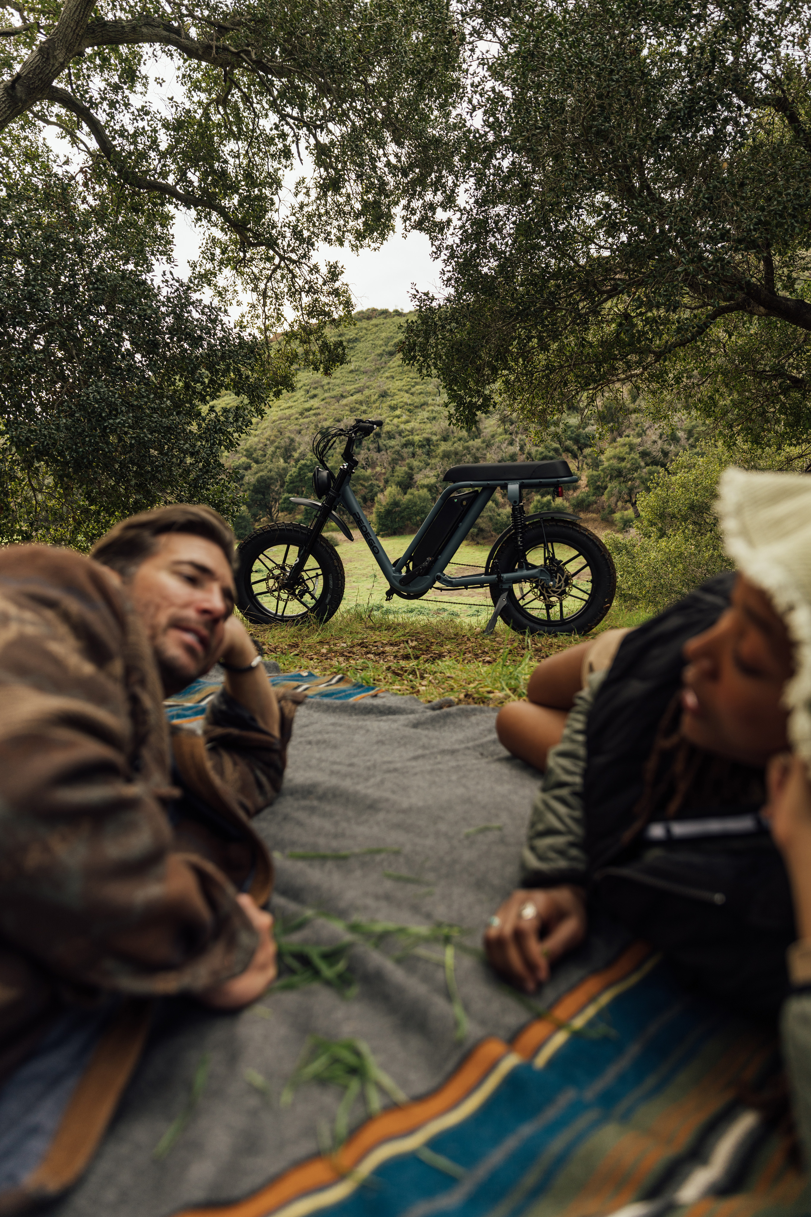 Two people lying on a picnic blanket under oak trees with a Pedego e-bike parked on the grass behind them