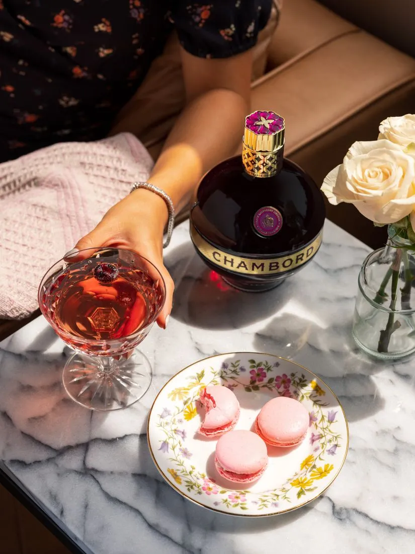 Person on a sofa holding a coupe of Chambord cocktail beside the bottle, macarons and roses on a marble table