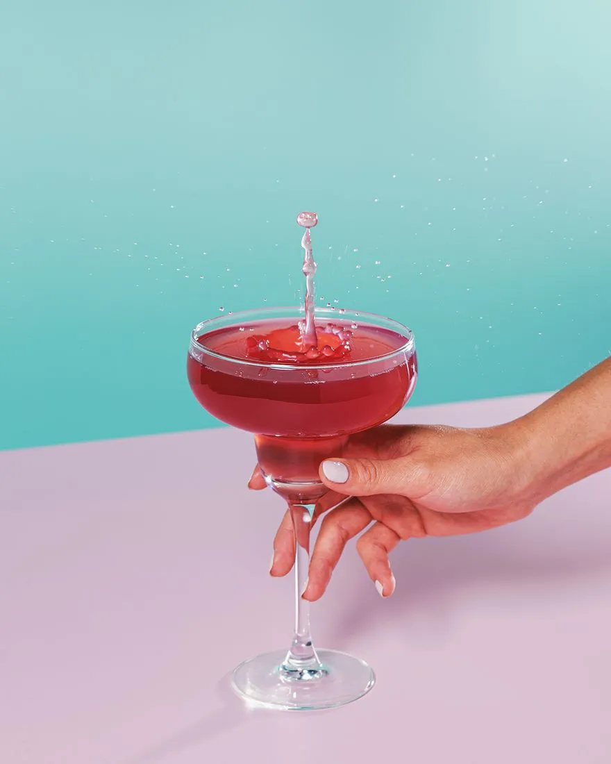 Hand holding a coupe of deep red Chambord cocktail mid-splash against a teal and pink background