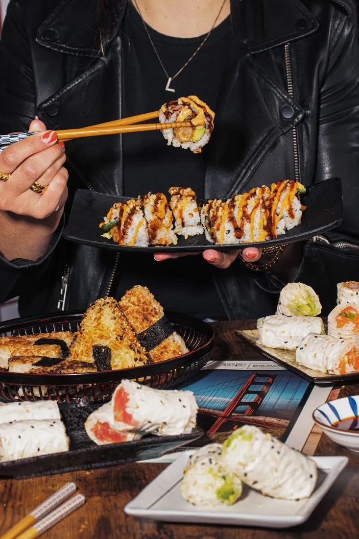 Person in a leather jacket lifting a piece of sushi with chopsticks over platters of rolls and tempura