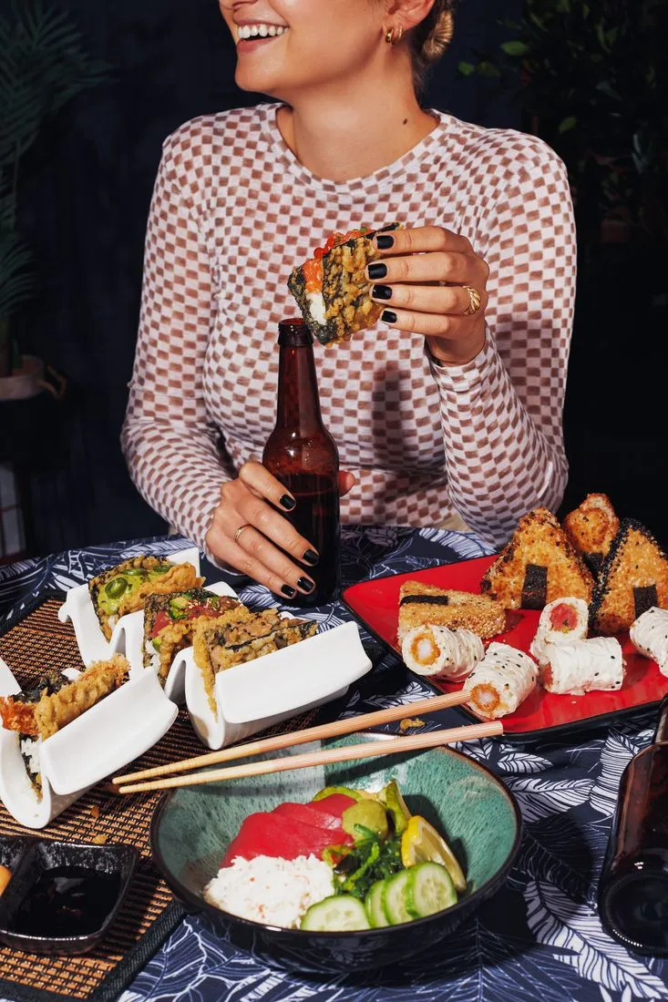 Person laughing at a table of sushi rolls, tempura and a poke bowl with a bottle of beer