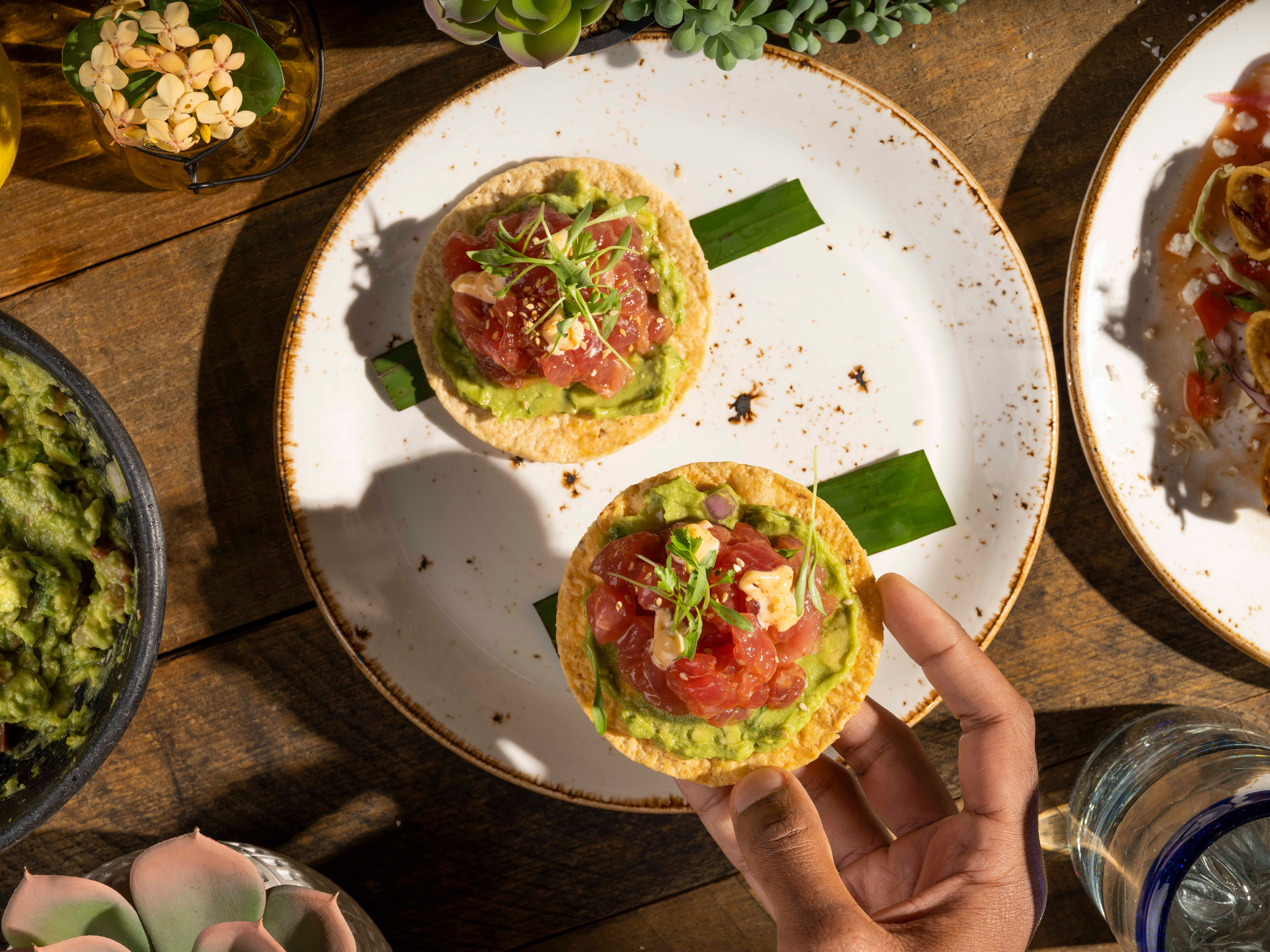 Overhead of tuna tostadas on avocado, held over a wooden table with guacamole and drinks