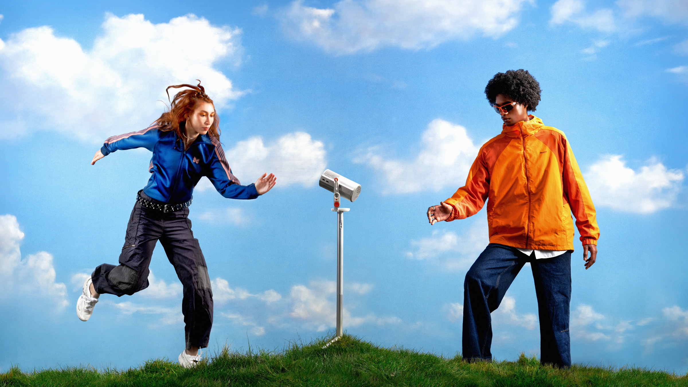 Two people dancing on a grassy hill either side of a projector on a stand, against a bright blue sky