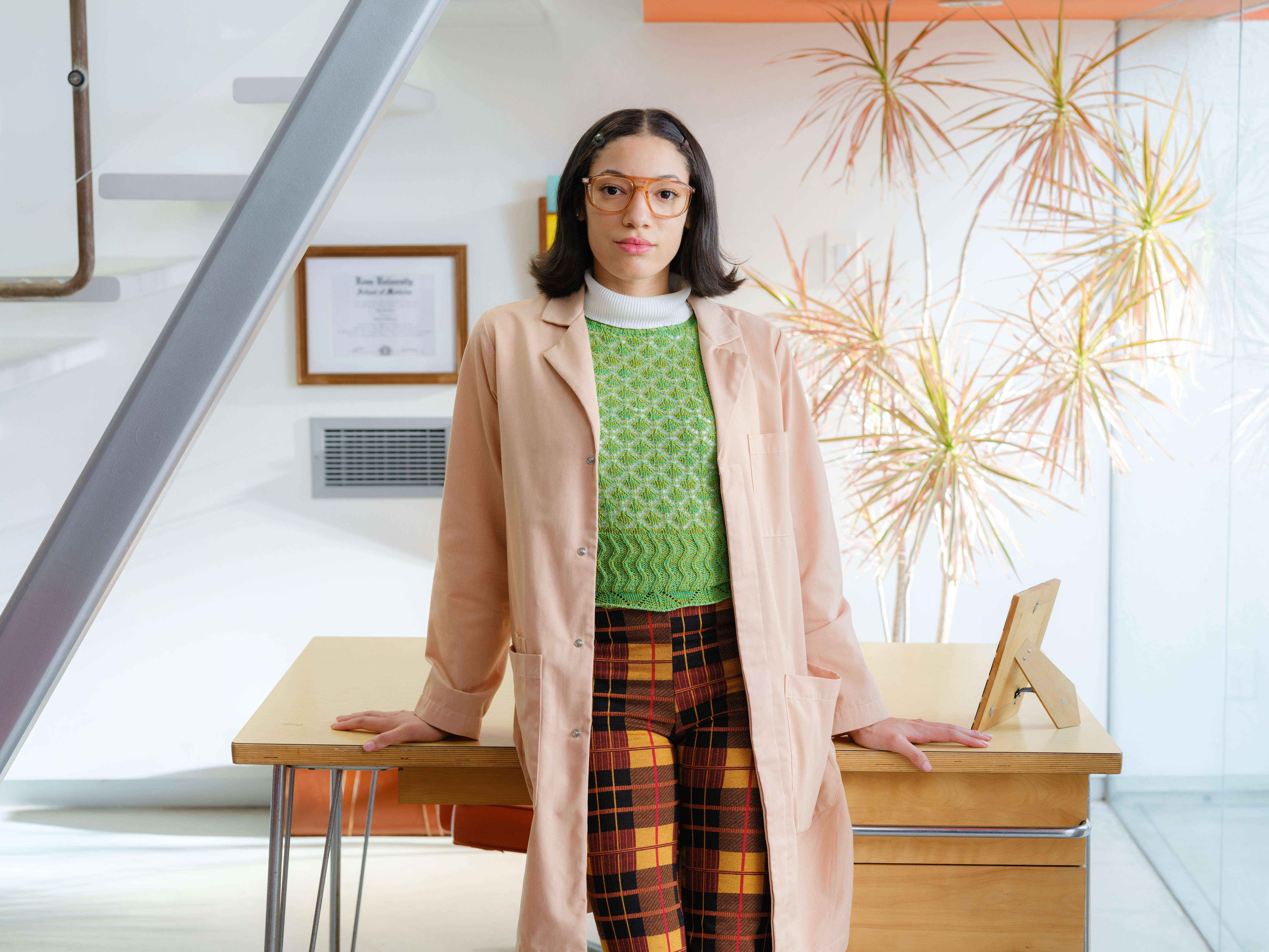 Person in a tan coat and Stoggles glasses standing at a desk in a bright modern office
